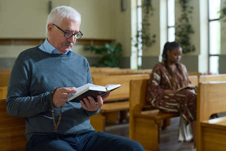 Serious mature man in casualwear looking through verses of Gospel while sitting on one of wooden benches during church serviceの写真素材