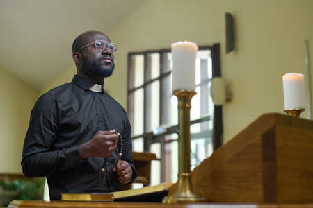 African American priest in black casualwear holding rosary beads during pray while standing by...