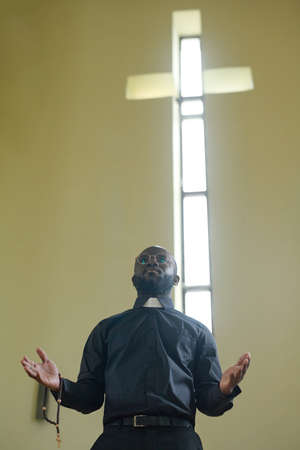 Young African American man in pastor apparel holding rosary beads while standing in front of parish...