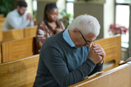 Mature man inclining his head and bending over his hands put together during silent pray after...