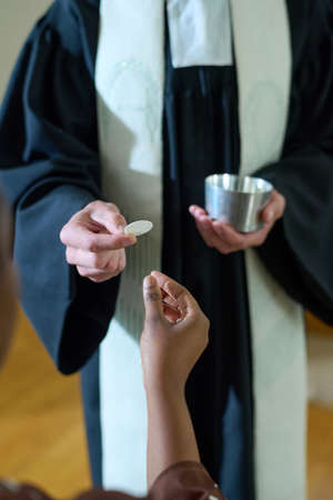 Hand of young Catholic priest in cassock passing unleavened bread to African American woman during communion rite in churchの写真素材