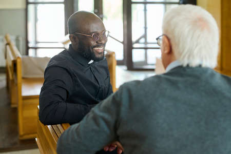 Young cheerful African American pastor of evangelical church consulting aged grey haired male parishioner after sermonの写真素材