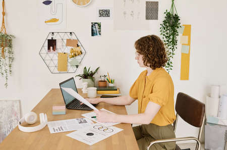 Side view of young female economist with financial papers using laptop while sitting by workplace...
