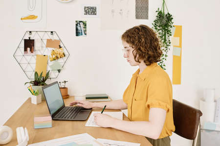 Young serious businesswoman in casualwear sitting by desk in front of laptop, making notes in financial document and analyzing dataの写真素材