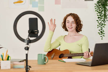Young cheerful woman waving hand to online audience during guitar playing lesson while sitting in...