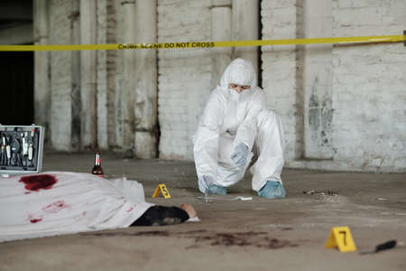 Criminological experts in coveralls, gloves and mask picking up evidences with metal pincers from asphalt while inspecting crime sceneの写真素材