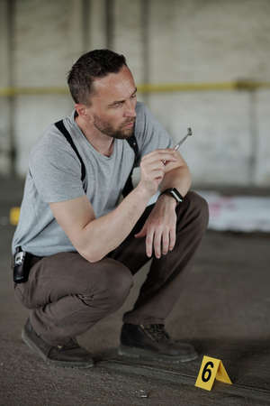 Detective inspector or policeman holding small evidence in metal pincers while sitting on squats in...