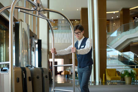 Young bellboy in uniform pushing cart with suitcases of hotel clients or guests while moving along corridor or crossing loungeの写真素材