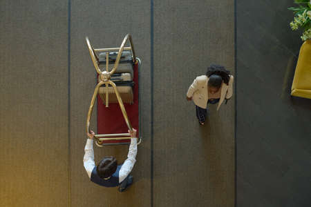Above view of young black woman in casualwear passing by bellboy pushing cart with baggage of guests...