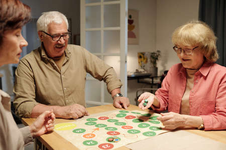 Happy mature woman throwing dice cubes over paper board with green and red circles of leisure game while playing with her friendsの写真素材