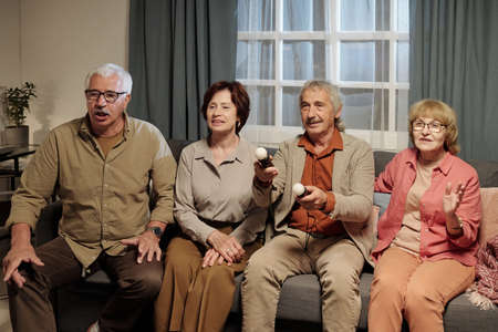 Four senior friends sitting in row on couch in front of tv set and playing leisure video game while...