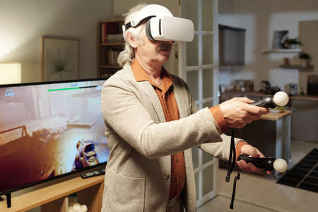 Senior man with vr headset controlling picture on tv screen with two gamepads while playing virtual game in living roomの写真素材