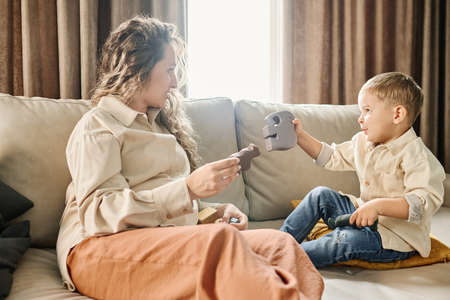Young woman with long blond wavy hair playing with toy animals with her cute little son and helping...
