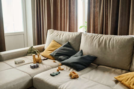 Wide leather couch of delicate grey color with large and small pillows and variety of toys in spacious cozy living room of modern apartmentの写真素材