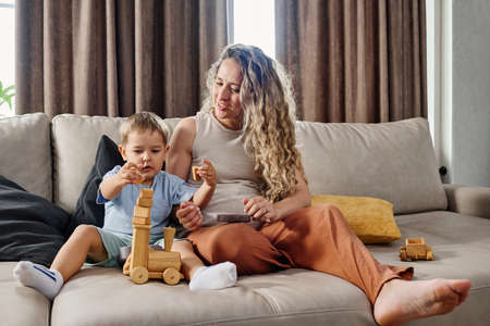 Cute little boy putting stack of wooden cubes on top of toy locomotive while sitting next to his mother on wide comfortable couchの写真素材