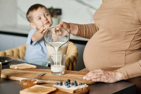 Close-up of young pregnant woman pouring fresh milk into glass for her cute little son sitting in...