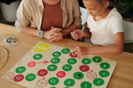 Youthful girl in whit et-shirt moving chip along paper with multi-color circles while playing board game with her mother by tableの写真素材
