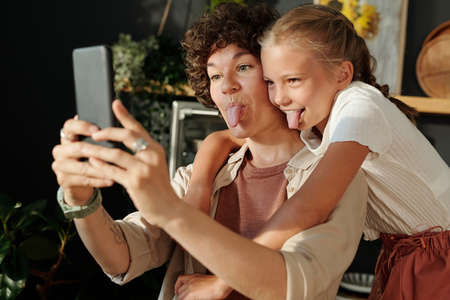 Cute youthful girl and her mother with smartphone showing tongues while making selfie or communicating in video chat at homeの写真素材