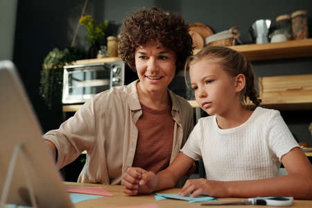 Young brunette woman pointing at tablet screen while sitting by table next to her daughter and...