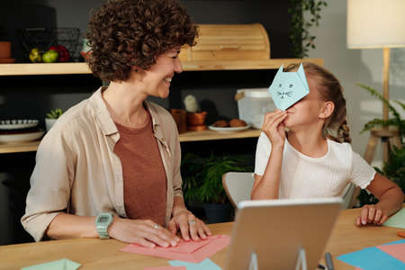 Cute girl holding paper cat mask close to her face while sitting by table in front of her mother and...