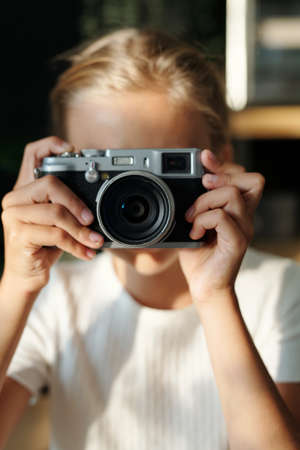 Hands of youthful blond girl holding retro photocamera by her face while taking shots of still life or people standing in front of herの写真素材