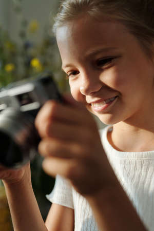 Smiling youthful girl with photo camera looking through new photos and enjoying shooting everything and everybody around herの写真素材