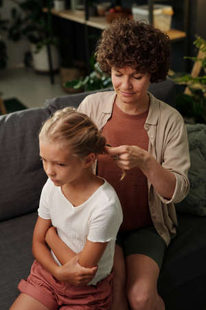 Young mother in casualwear plaiting long blond hair of her cute youthful daughter while both sitting on couch in living roomの写真素材