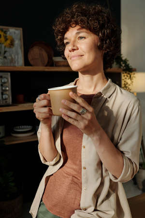 Young smiling brunette with short dark curly hair holding mug with tea or coffee while standing in front of window in the morningの写真素材