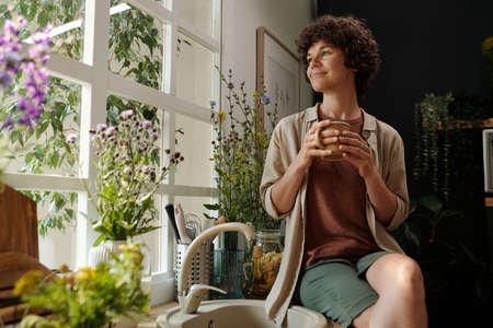 Young serene brunette with cup of coffee looking through window surrounded by plants while sitting...