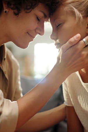Happy young woman touching her cute youthful daughter by hand and forehead while sitting in front of her and slightly bending forwardsの写真素材