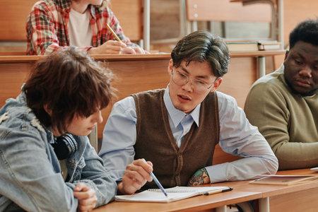 Asian guy in smart casualwear explaining his girlfriend notes in copybook while sitting next to her by desk against intercultural classmatesの写真素材
