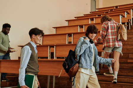 Group of intercultural teenage students in casualwear having talk while entering lecture hall with desks before seminar or lessonの写真素材