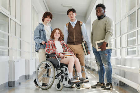 Group of multicultural teenage students in casualwear standing in college corridor while one of them sitting in wheelchairの写真素材