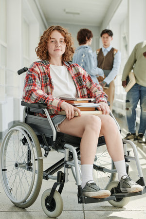 Cute teenage female student in wheelchair sitting in front of camera in college corridor against group of her classmates talking at breakの写真素材