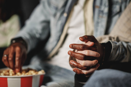 Selective focsu shot of unrecognizable Black man and Caucasian woman in love watching movie at cinema holding handsの写真素材