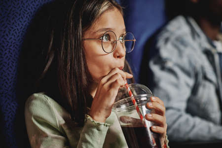 Joyful preteen girl wearing eyeglasses sipping carbonated sweet drink while watching movie at cinemaの写真素材