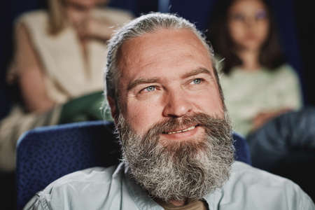 Horizontal close-up portrait of joyful mature Caucasian man with beard on face watching comedy movie at cinemaの写真素材