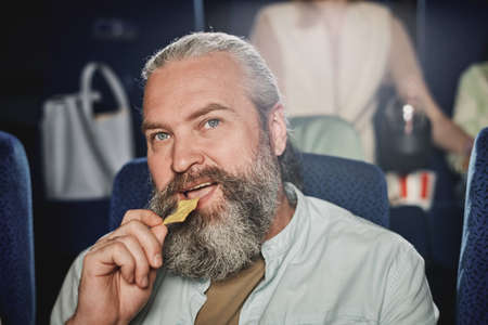 Close-up of mature Caucasian man with beard on face enjoying having snack and watching film at cinemaの写真素材