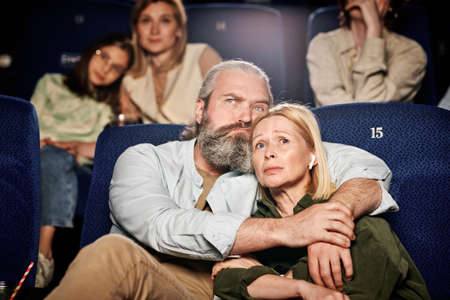 Mature adult Caucasian man with beard on face hugging his wife while watching sad drama film at...