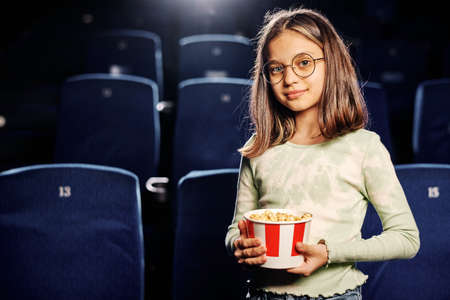 Horizontal medium portrait of beautiful preteen Caucasian girl holding bucket of popcorn standing in cinema hall against navy blue seatsの写真素材