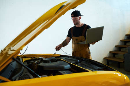 Young mechanic with laptop installing wires into engine of car while carrying out technical diagnostics of motor after repairmentの写真素材