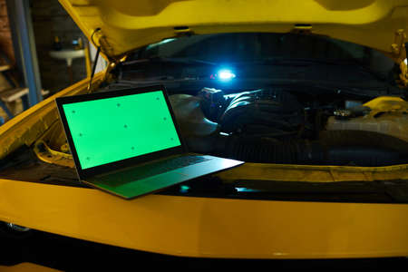 Laptop with green screen standing on engine of electric car of yellow color with open hood before or during technical checkupの写真素材