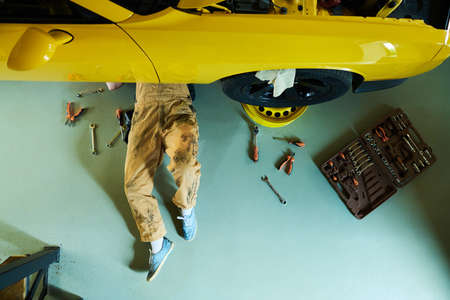 Above view of repairman in overalls and gumshoes lying under yellow car on the floor of workshop or...