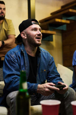 Happy young man in denim jacket and cap pressing buttons of gamepad while sitting on couch in front...