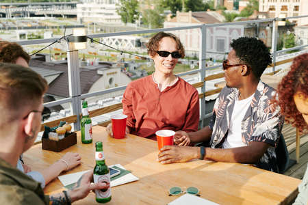 Young cheerful interracial men and their girlfriends in stylish casualwear chatting by table in...