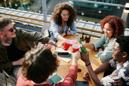 Five cheerful youthful friends clinking with their drinks over table while having rest in outdoor...
