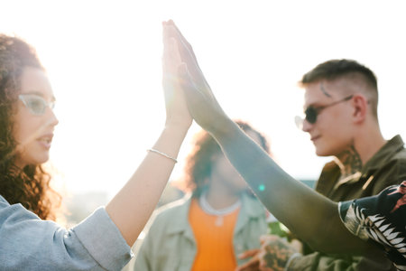 Young intercultural dates giving each other high five against their friends having chat during outdoor party in rooftop cafe or restaurantの写真素材