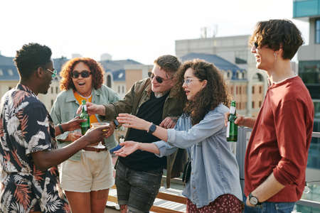 Young black man passing bottle of beer and cocktail to guy and girl during rooftop party or...