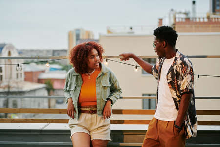 Young African American couple in stylish casualwear having chat on terrace of rooftop cafe against urban environmentの写真素材