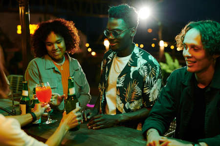 Group of young intercultural friends taking ordered cocktails and beer while standing by counter and...